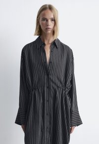 Schwarz-weiße gestreifte Hemdbluse mit Knopfleiste, Kragen und elastischem Bund. Verfügt über lange Ärmel mit breiten Manschetten.