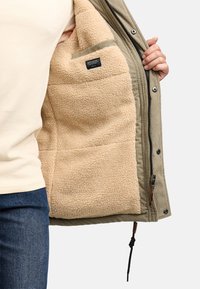 Eine Person in blauen Jeans und einem beigefarbenen Hemd hält eine khakifarbene Jacke auf, die mit einem beigen Fleece-Futter und einer Innentasche mit Etikett ausgestattet ist.