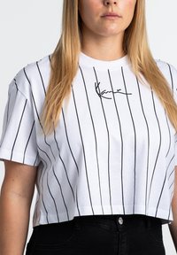 Weißes kurz geschnittenes T-Shirt mit schwarzen vertikalen Streifen und besticktem Signaturdetail auf der Brust. Weiches Baumwollmaterial, lockere Passform.