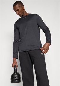 Langärmliges schwarzes Sportshirt und passende Hose, beide aus glattem Stoff. Das Shirt hat ein kleines weißes Logo auf der Brust.