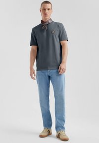 Grijze katoenen polo met een logo op de borst, gecombineerd met lichtblauwe jeans en beige schoenen met zwarte accenten, aangevuld met een gebreide sjaal.