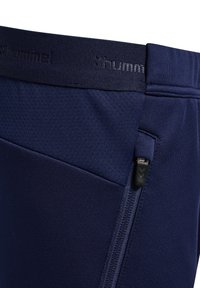 Marineblaue Sportshorts aus strukturiertem Material, mit elastischem Bund und einer seitlichen Reißverschlusstasche mit schwarzem Beschlag. Hummel-Logo am Bund.