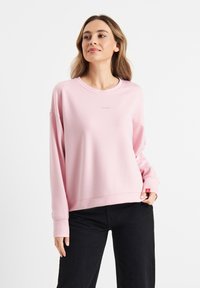 Light pink sweatshirt with a round neckline, dropped shoulders, ribbed cuffs, and a small logo printed on the chest. Paired with black pants.
