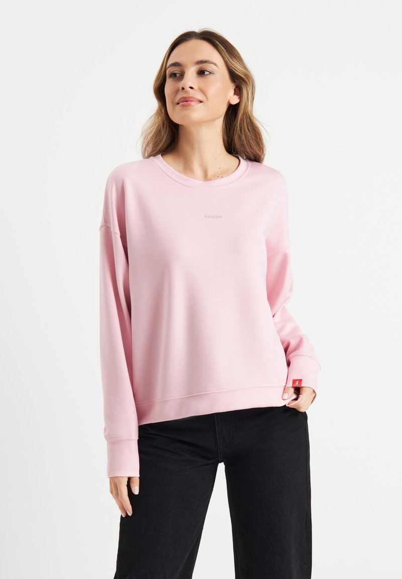 Light pink sweatshirt with a round neckline, dropped shoulders, ribbed cuffs, and a small logo printed on the chest. Paired with black pants.