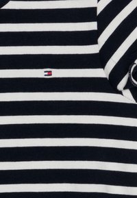 Tessuto a maglia a righe blu navy e bianche con un piccolo logo rosso, bianco e blu sul petto. Collo rotondo con maniche corte.