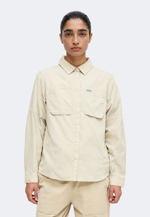 Femme aux cheveux foncés portant une chemise beige boutonnée et un pantalon assorti, debout face à l'avant avec une expression neutre sur un fond uni.