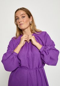 Lila Kleid mit langen, aufgepufften Ärmeln und einem Bindegürtel; strukturierter Stoff mit dezenten Falten am Ausschnitt; goldene Knopfakzente an den Ärmeln.