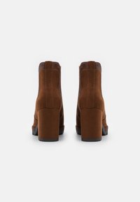 Un paio di stivaletti alla caviglia in suede marrone con tacco a blocco e accenti in pelle marrone scuro nella parte superiore, mostrati da dietro su uno sfondo bianco.