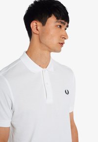 Weißes Polohemd aus strukturiertem Stoff, mit einem schwarzen gestickten Logo auf der Brust und einem klassischen Kragen mit Knopfverschluss.