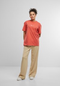 Korálová bavlnená tričko s voľným strihom a krátkymi rukávmi, s bielym logom. V kombinácii s béžovými širokými nohavicami a bielymi športovými teniskami.