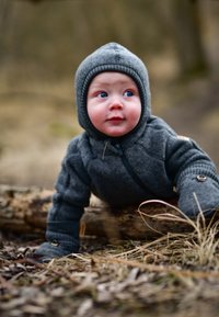 Grauer Wollanzug mit Kapuze, ausgestattet mit einem Reißverschluss und strukturierten Bündchen, getragen von einem krabbelnden Kind auf einem Baumstamm umgeben von trockenem Gras.