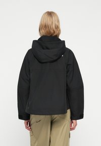 Oseba s ramenskimi lasmi v blond barvi, oblečena v črno kapuco jakno in bež cargo hlače, prikazana s hrbtne strani na enobarvni ozadju.