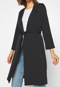 Trench-coat noir à manches longues avec ceinture à nouer à la taille, revers ajustés et poches latérales. Texture lisse avec une coupe décontractée, porté avec un jean.