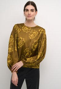 Arany selyem blúz nagy sárga virágmintával, kerek nyakkivágással, hosszú puffos ujakkal, melyek a csuklónál vannak megkötve, fekete nadrággal párosítva.