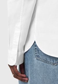 Camicetta bianca a maniche lunghe con polsino abbottonato, caratterizzata da una texture liscia, orlo a coda di rondine arrotondato e abbinata a jeans in denim azzurro chiaro.
