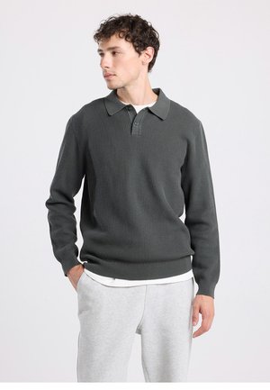 Donkergroene, gebreide trui met een kraag, lange mouwen en een geribbelde structuur. Draagt over een wit overhemd, gecombineerd met grijze joggingbroek.