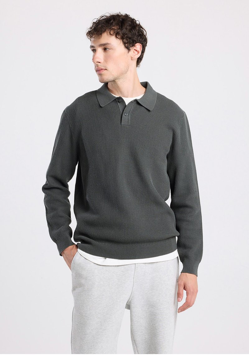 Tummanvihreä, neulottu villapaita, jossa kauluksellinen kaula-aukko, pitkät hihat ja resorakenteinen pinta. Päällä valkoisen paidan päällä, yhdistetty harmaisiin collegehousuihin.