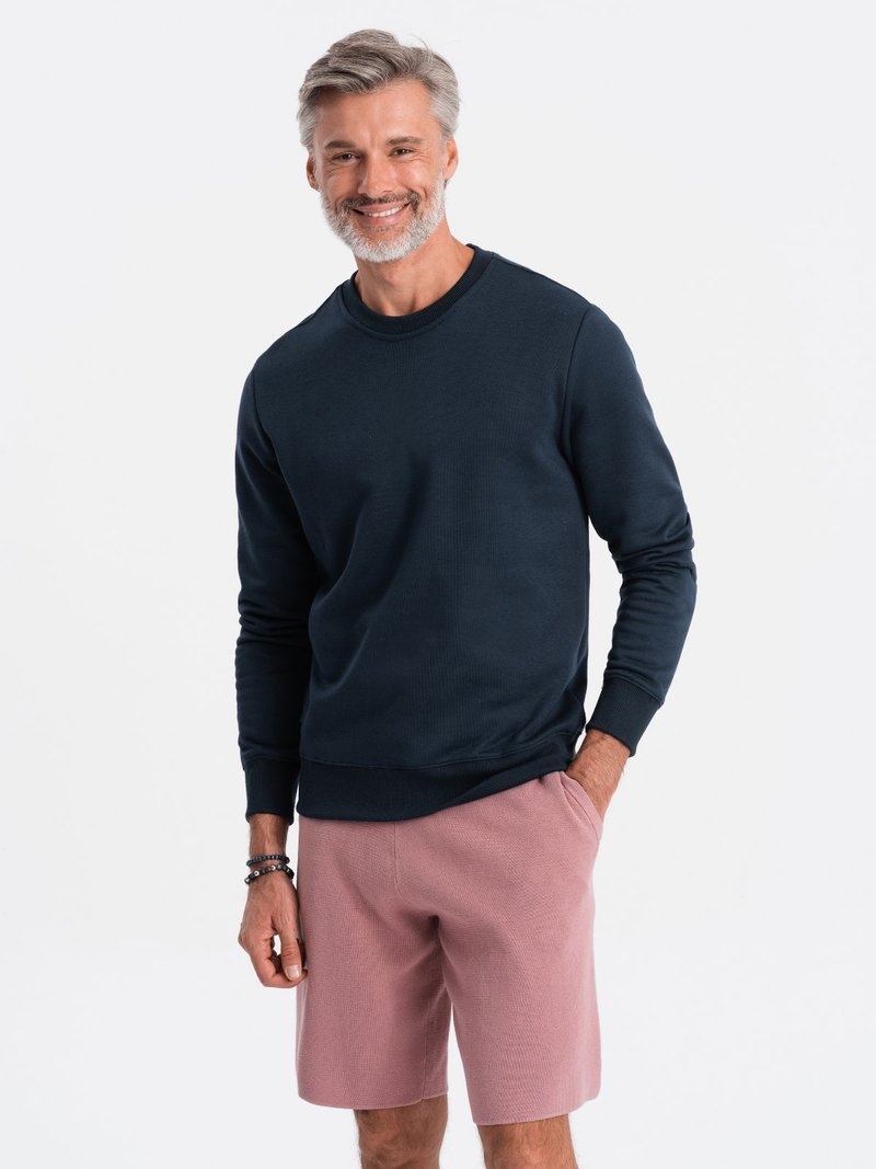 Marineblauwe sweatshirts met ronde hals en geribde mouwen, gecombineerd met lichtroze textuurs shorts. Model staat met een lichte glimlach.
