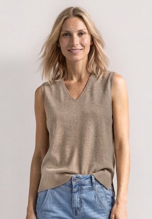 Femme souriante aux cheveux blonds mi-longs portant un top beige en tricot sans manches et un jean bleu clair sur un fond uni.