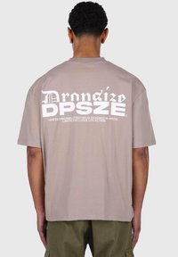 Braunes Baumwoll-T-Shirt mit großen weißen Buchstaben auf dem Rücken, kurzen Ärmeln und einem lockeren Schnitt. Der Saum ist gerade und schlicht.