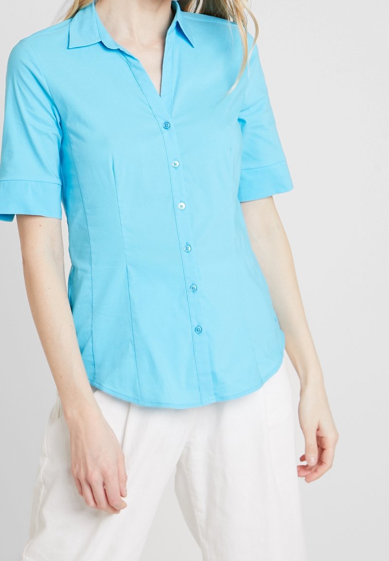Femme portant une chemise cintrée bleu clair à manches courtes boutonnée et un pantalon blanc ample sur un fond uni.