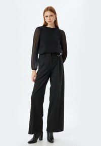 Blusa nera in chiffon con maniche a sbuffo, abbinata a pantaloni a gamba larga a righe con vita alta e stivaletti neri con tacco.