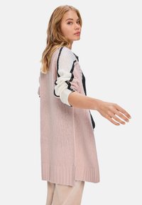 Hellrosa gestrickte Strickjacke mit langen Ärmeln, ausgestattet mit Schwarz-weiß-Akzenten. Strukturiertes Material und gerippter Saum. Offenes Design, über einem dunklen Oberteil getragen.