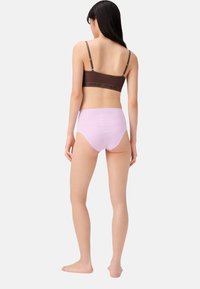 Donna in piedi a piedi nudi, girata di spalle, che indossa un reggiseno marrone a croce con spalline regolabili e mutandine viola a vita alta.