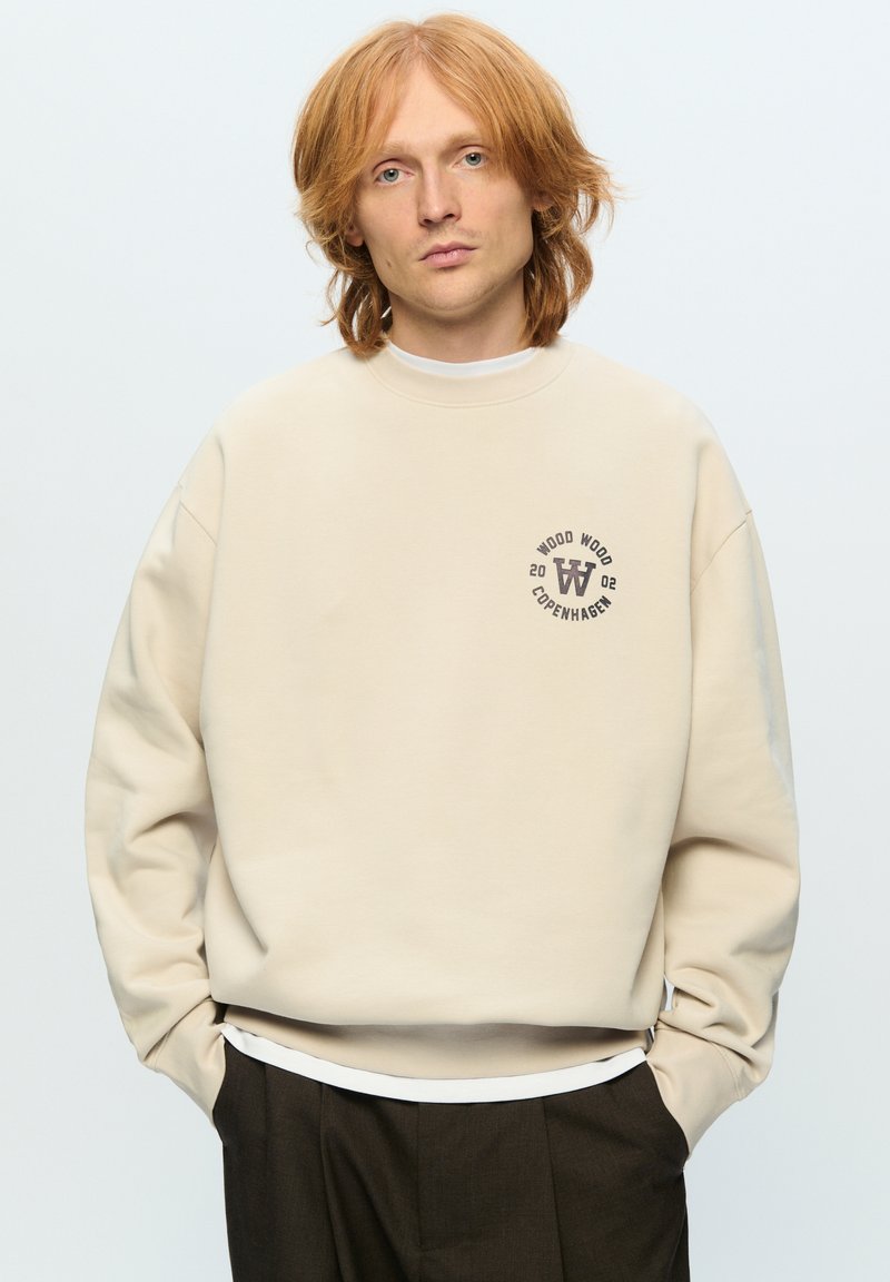Beige sweatshirt met een ronde hals en een klein rond logo op de borst. De stof lijkt zacht te zijn, met een ontspannen pasvorm.