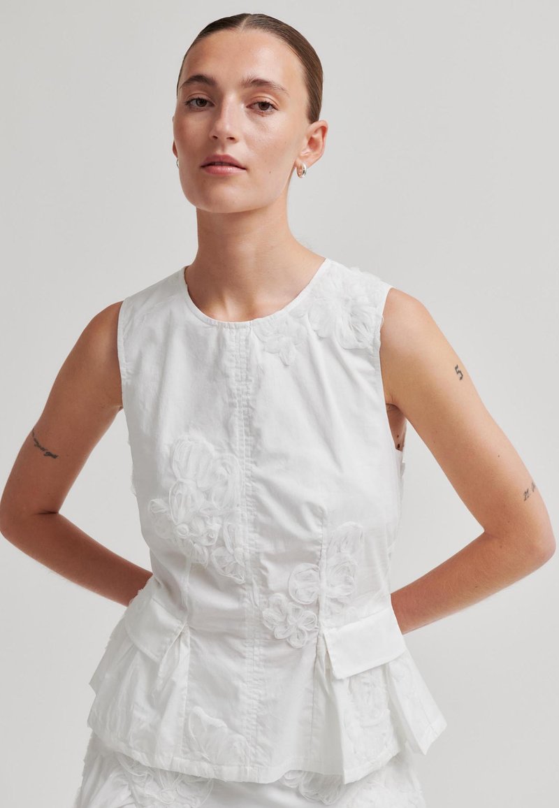 Femme portant un chemisier blanc sans manches avec broderie florale et détail péplum, debout les mains derrière le dos sur un fond uni.
