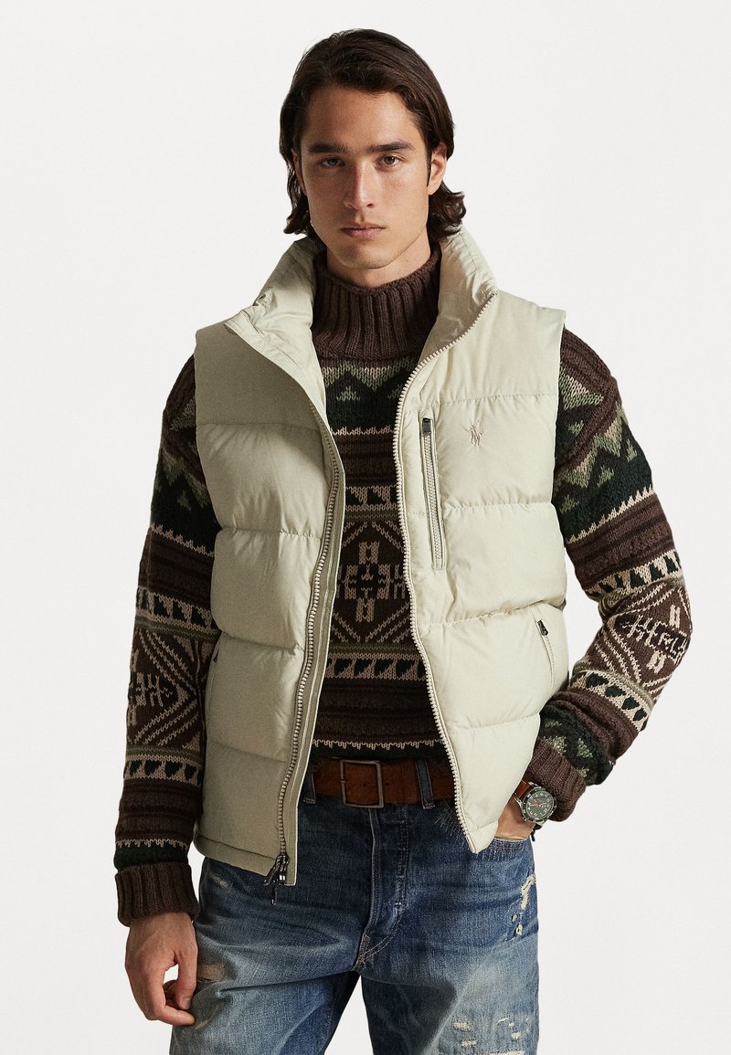 Colete puff de tom bege claro, com gola alta, fecho de zipper e bolsos laterais, usado sobre um suéter de malha castanho com padrão e jeans de denim.