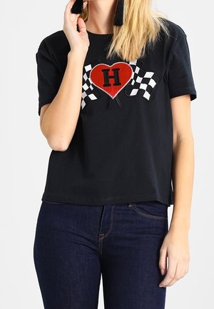 Nainen pukeutunut mustaan t-paitaan, jossa on punainen sydän ja kirjain H kahden mustavalkoruudullisen lipun välissä, yhdistetty tummansinisiin farkkuihin.