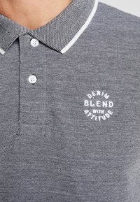 Grå polo skjorte med strukturert stoff, hvite detaljer på kragen, og en sirkulær logo med teksten "DENIM BLEND med ATTITUDE."