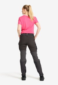 Kvinna med blont hästsvans som står med händerna på höfterna, iförd en klarrosa kortärmad skjorta och svart-grå cargo-byxor med svarta skor.