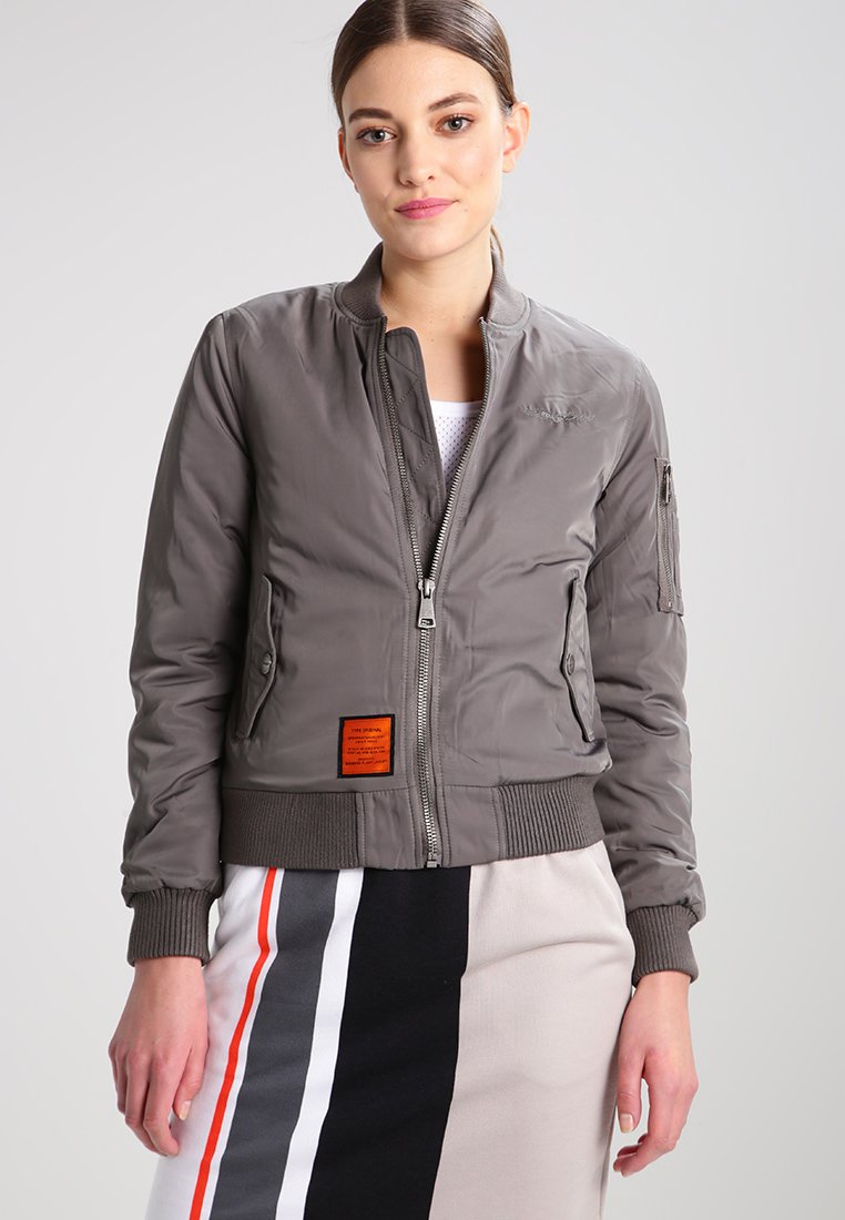 Graue Bomberjacke mit einer leichten, glatten Textur. Hat einen Frontreißverschluss, gerippte Bündchen und ein oranges Etikett auf der linken Seite.