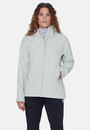 Kvinde med krøllet hår iført lysegrå hættetrøje og mørke bukser, stående med den ene hånd i jakkelommen.