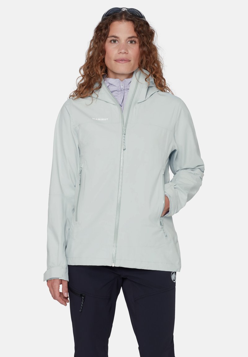 Γυναίκα με σγουρά μαλλιά που φοράει ανοιχτό γκρι μπουφάν με κουκούλα και σκούρο παντελόνι, στέκεται με το ένα χέρι στην τσέπη του μπουφάν.