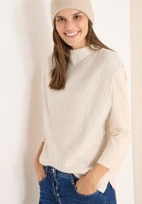 Beige Rippstrickpullover mit hohem Kragen, über einem hellen Langarmtop getragen, kombiniert mit blauen Jeans. Getragen mit einem passenden Hut.