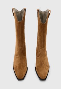 Botas de ante marrón hasta el tobillo con punta afilada, diseño texturizado y pequeñas aberturas laterales. Suela negra con detalles sutiles en la parte superior.