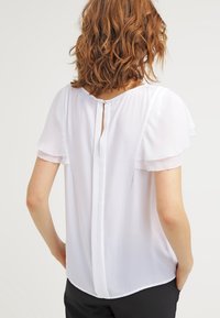 Bílá blůza s krátkými volánkovými rukávy, detaily na zádech ve tvaru klíčové dírky a volným střihem. Vyrobena z lehké, poloprůhledné tkaniny.