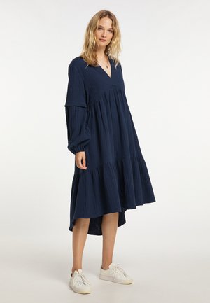 Blonde Frau trägt ein lockeres, langärmeliges marineblaues Midikleid mit weißen Sneakers und steht vor einem schlichten weißen Hintergrund.