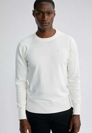 Homme portant un pull col rond blanc ajusté à manches longues avec un petit logo sur la poitrine et un pantalon noir, debout devant un fond gris uni.