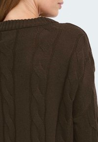 Brun kabelstrikket genser med ribbestrikket hals og teksturerte mønstre på kroppen. Stoffet ser mykt og tykt ut.