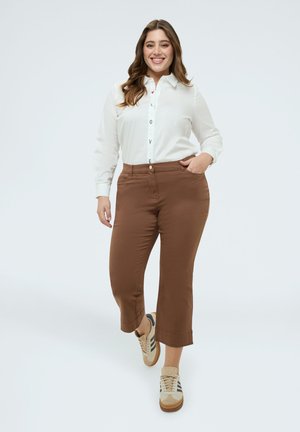 Smilende kvinde, der går fremad iført en hvid bluse med "LOVE" knapper, brune cropped bukser og beige sneakers med sorte striber.