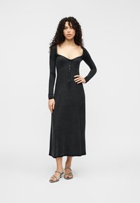 Μαύρο, εφαρμοστό φόρεμα με μακριά μανίκια, τετράγωνο ντεκολτέ, κλείσιμο με κουμπιά μπροστά και φουσκωτή λαιμόκοψη. Φορεμένο με σανδάλια με λουράκια πάνω σε ουδέτερο φόντο.