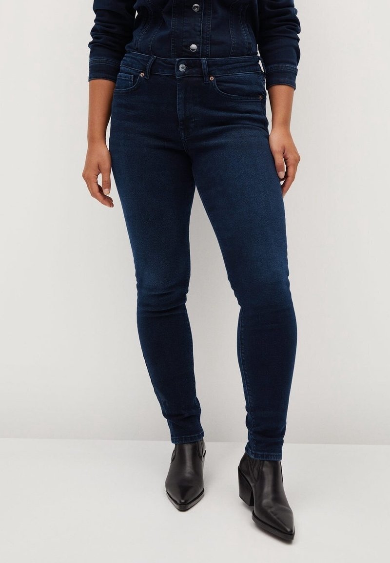 Persona que lleva jeans ajustados de tiro alto de color azul oscuro y botines negros de punta puntiaguda, de pie frente a un fondo liso y claro.