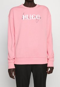 Personne portant un sweat-shirt à manches longues rose avec le logo « HUGO » en texte blanc et noir cursif sur la poitrine, associé à un pantalon noir.