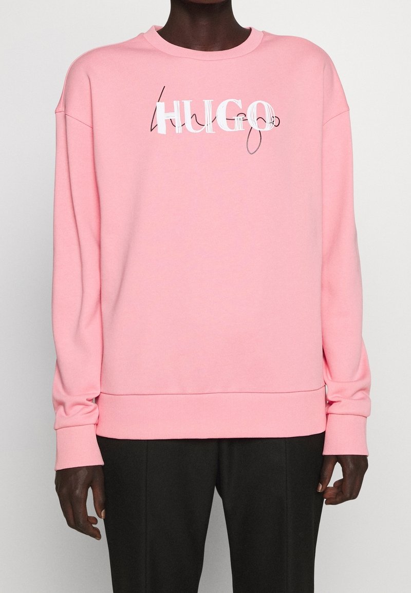 Personne portant un sweat-shirt à manches longues rose avec le logo « HUGO » en texte blanc et noir cursif sur la poitrine, associé à un pantalon noir.