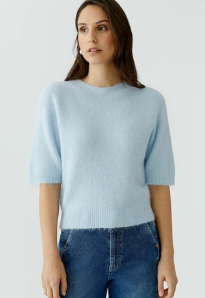 Vrouw staand, dragend een lichtblauwe, zachte trui met korte mouwen en een blauwe spijkerbroek, licht naar links kijkend tegen een effen achtergrond.