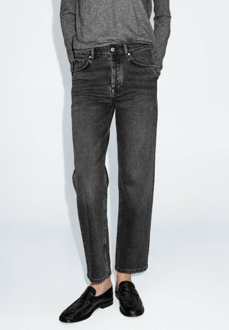Jeans in denim nero con una vestibilità dritta, dotati di chiusura con bottoni, design a cinque tasche e dettagli di cucitura sottili. Indossati con mocassini neri.